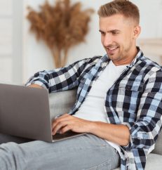 Freelance And Remote Work Concept. Portrait of positive man in checkered shirt chatting online using laptop, sitting on sofa in cozy living room. Smiling freelancer working on computer at home