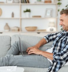 Side view of young happy man chatting online on laptop with friend at home, lying on couch in light livingroom, copyspace