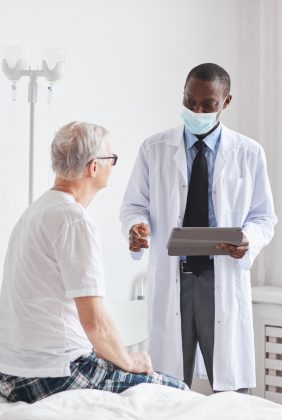 Portrait of African-American doctor talking to senior man in white hospital room, copy space