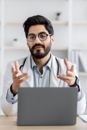 Video call consulting with doctor. Private clinic support. Friendly millennial arabian bearded guy in white coat, glasses with stethoscope at table with laptop, gesturing in office in clinic interior